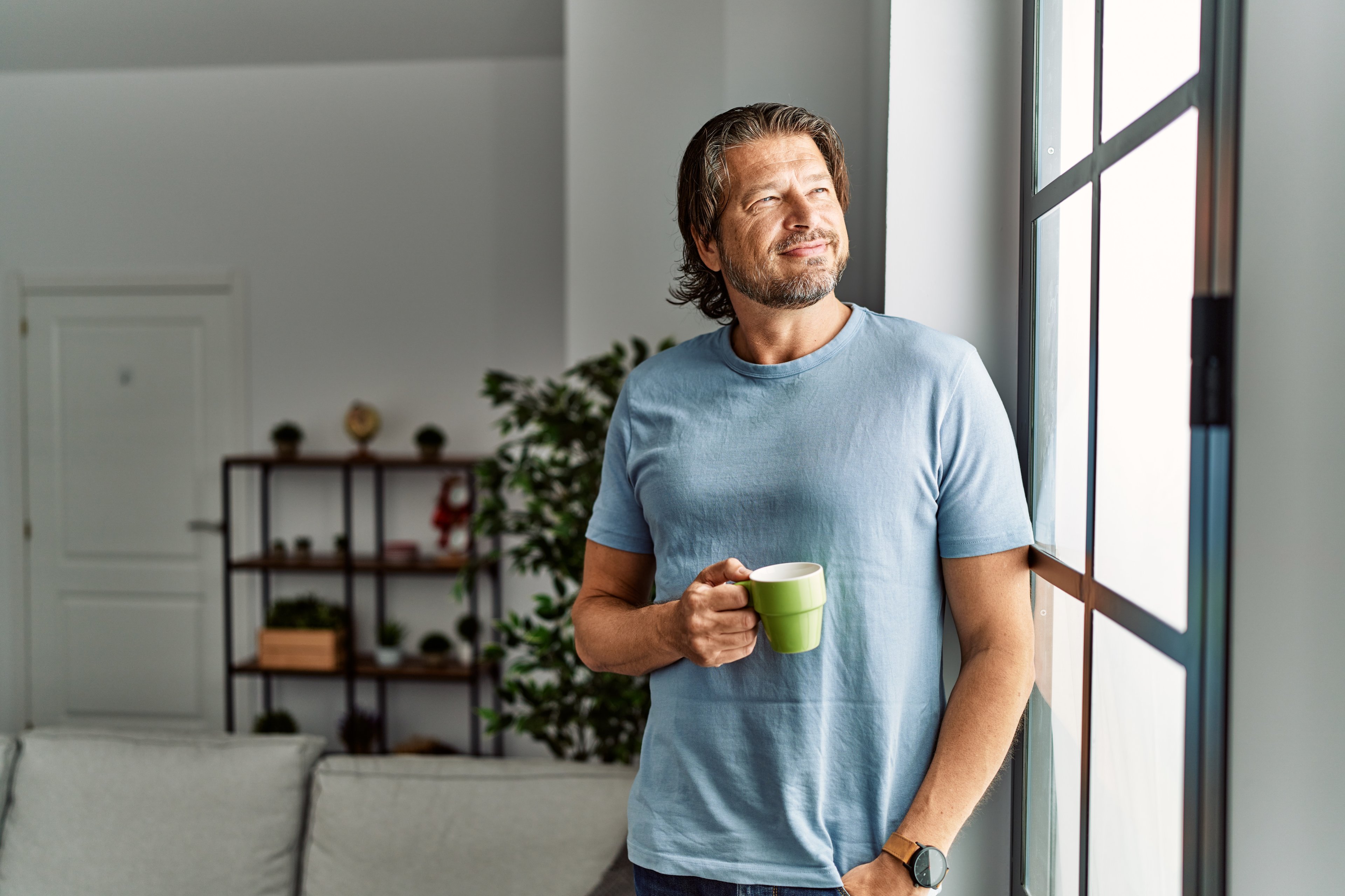Middle-aged man looking out a window with a cup of coffee in his hands. 