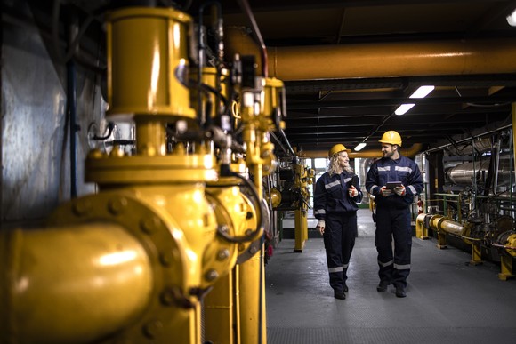 Two engineers walk through an industrial control room full of yellow pipes. 