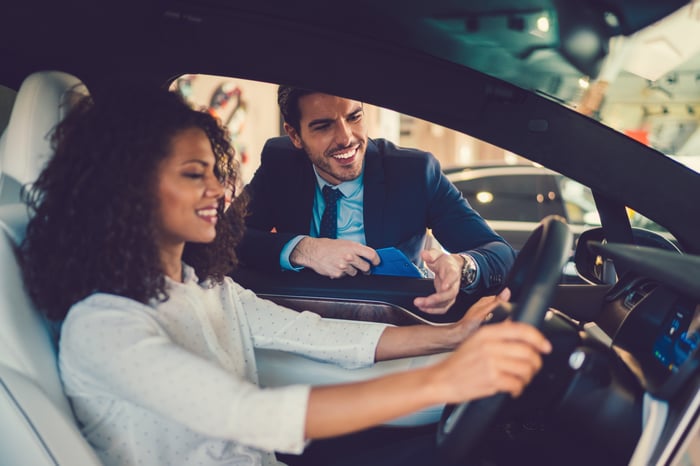 Person behind wheel of car, with another person looking in window.