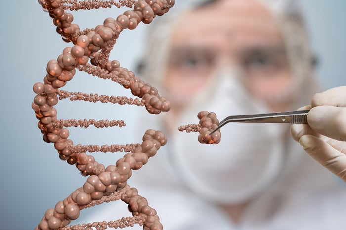 A scientist in the background wearing a respirator and holding forceps appears to be removing a piece of a DNA model in the foreground.