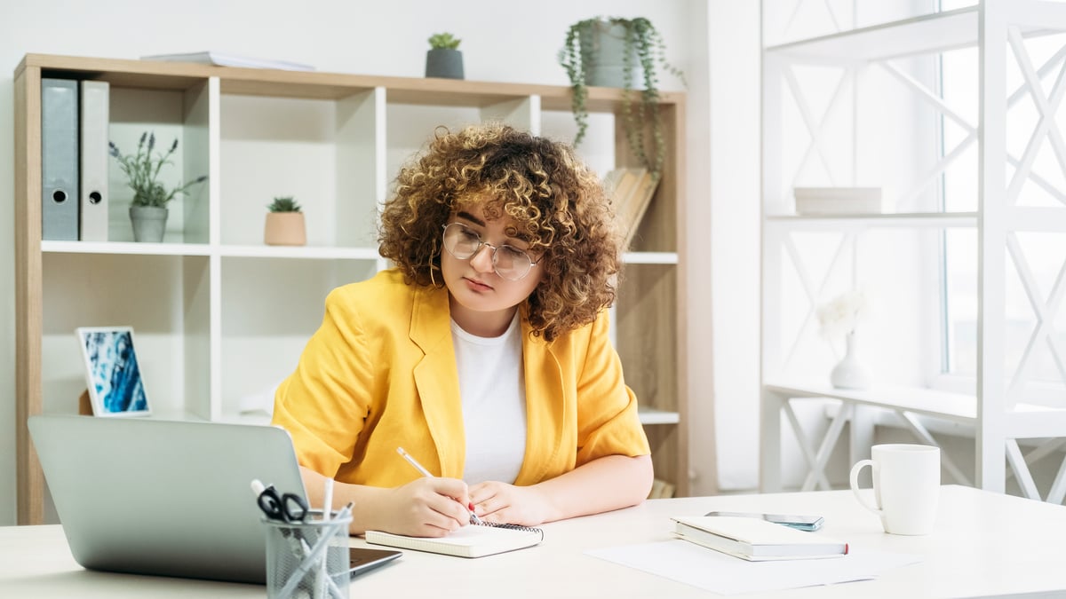 A person writing in a notepad while looking at a laptop.