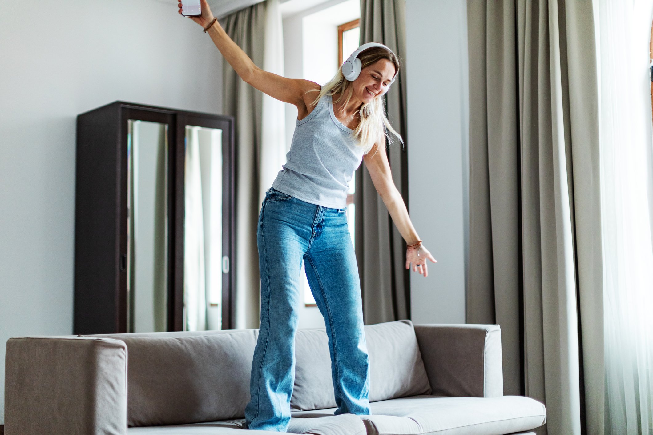 A woman in jeans and a tank top wearing headphones and dancing on her sofa.
