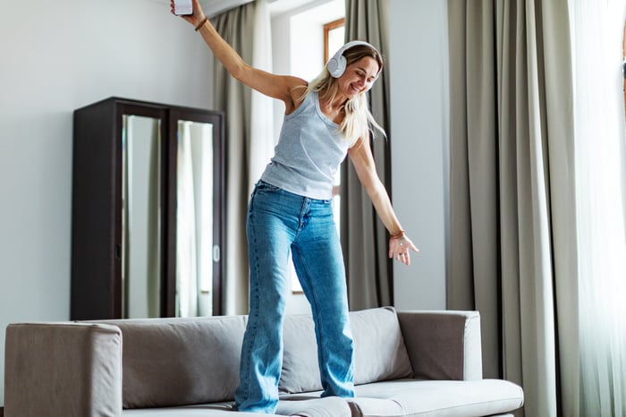 A woman in jeans and a tank top wearing headphones and dancing on her sofa.