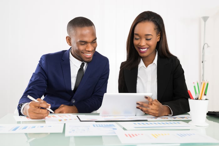 Two business people looking at a tablet on a desktop covered with stock charts. 