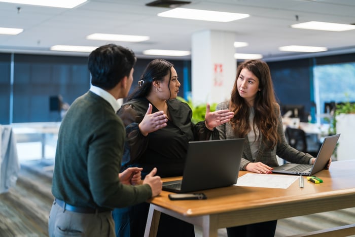 Three people having a discussion at a table with computers on it.