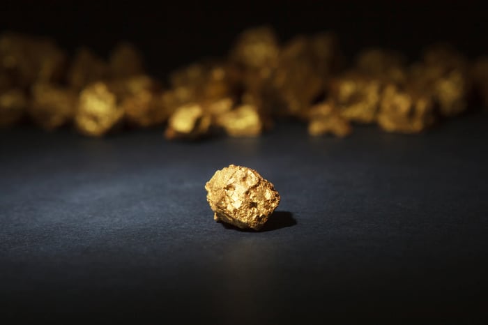 A close-up photograph of gold nuggets. A single nugget is in the foreground, while others are blurred out in the background. 
