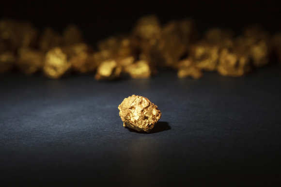 A close-up photograph of gold nuggets. A single nugget is in the foreground, while others are blurred out in the background. 