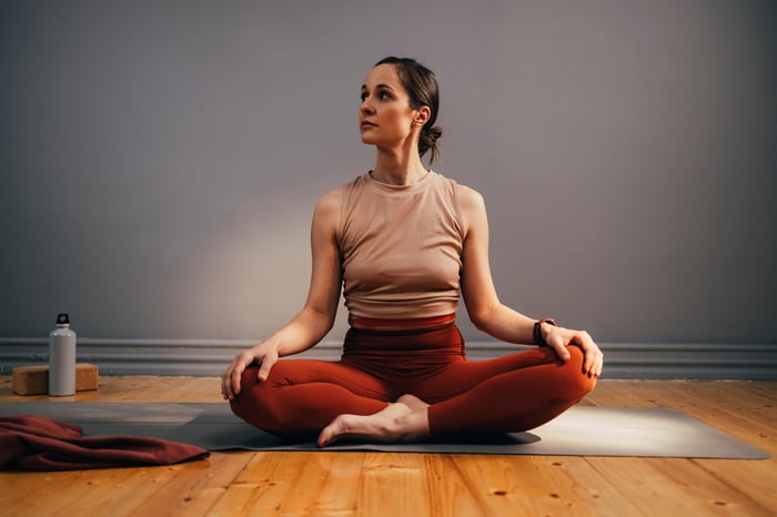 A woman sitting on a yoga mat cross legged looking to the side. 