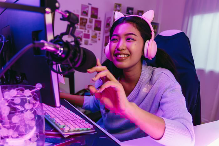 A smiling person with microphone, headphones, and pink cat ears next to computer. 