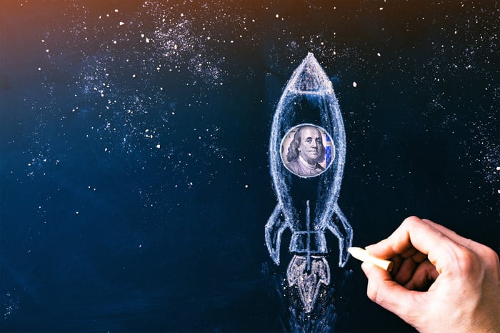 A hand holding a piece of chalk next to a drawing of a rocket with a picture of Ben Franklin from a $100 bill peering through a window on the rocket.