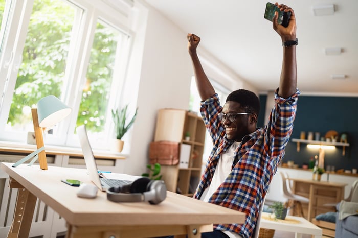 A smiling person holding their hands up in the air and looking at a laptop.