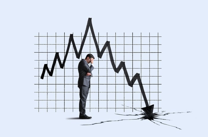 A person with their head in their hand stands in front of a stock chart on which an arrow is crashing into the ground.