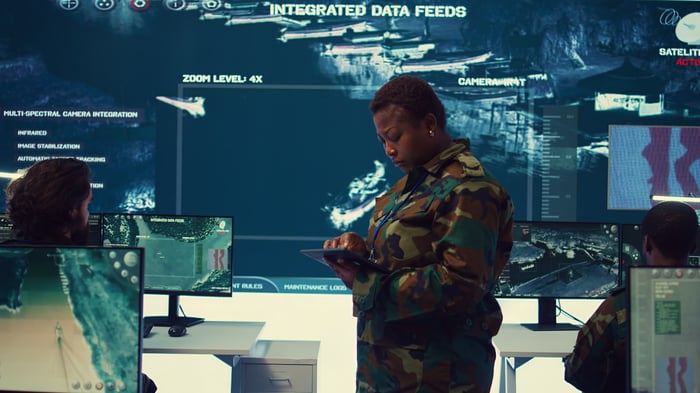 Two U.S. Army soldiers in some sort of control room with wall-mounted video screens showing data.