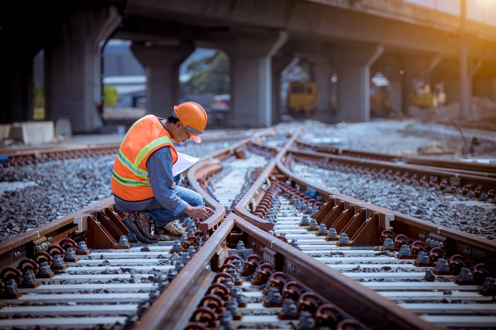 A worker wearing a hard hat kneeling at a junction of railroad tracks.