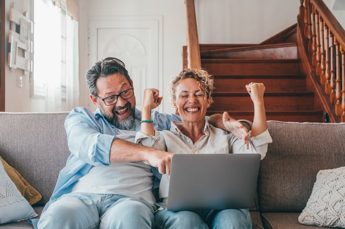 A couple is on a couch, smiling and laughing at an open laptop, and pumping fists.