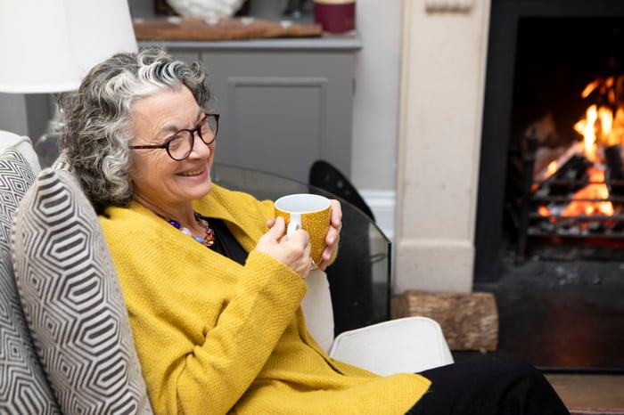 Someone is sitting by a fire with a hot drink, smiling.