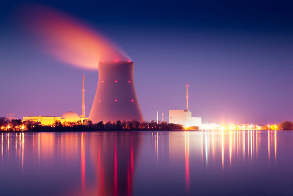 A nuclear power plant at night, with a cooling tower emitting steam and lights reflecting on a body of water. 