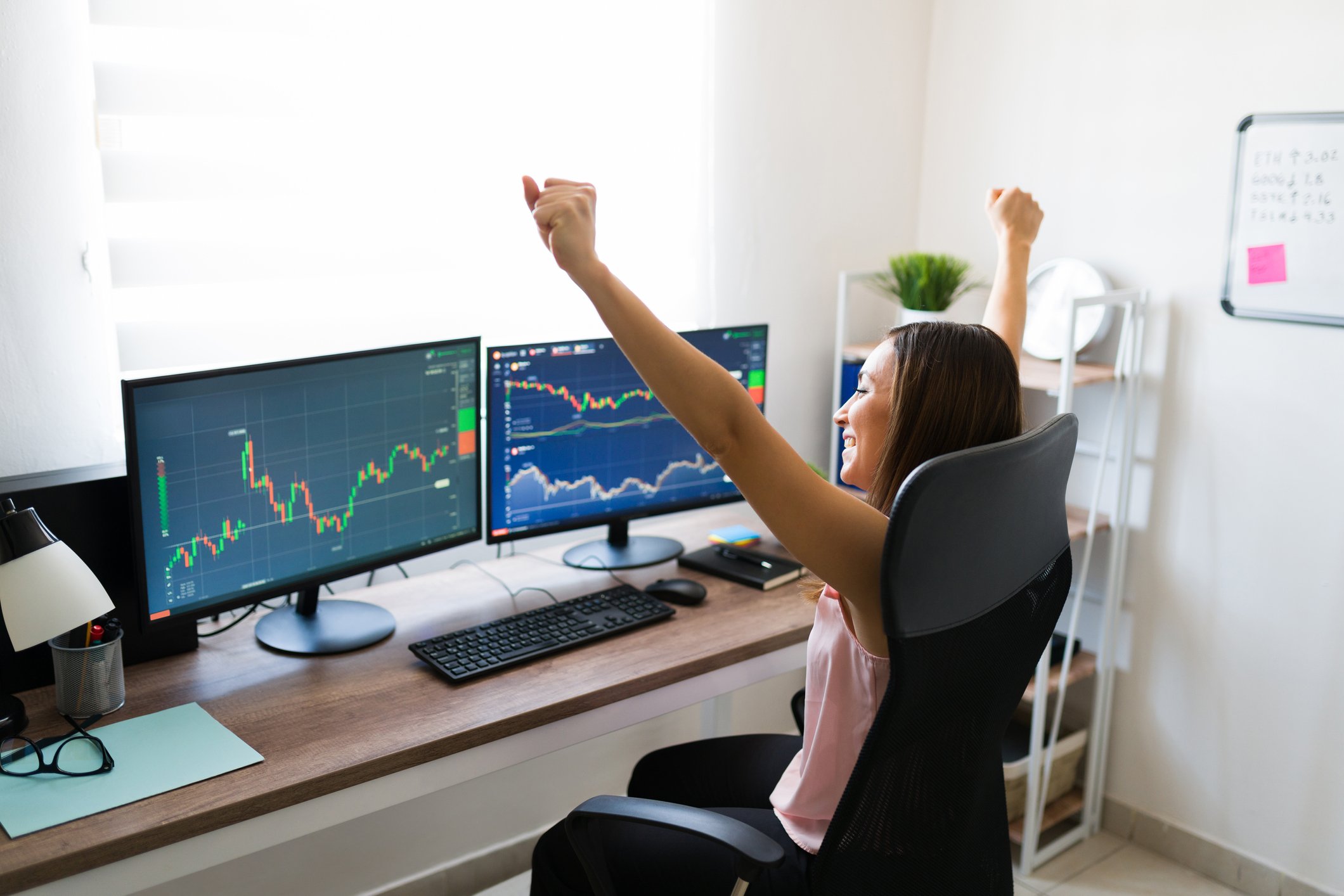 A person holding hands up in the air while looking at monitors displaying stock charts.