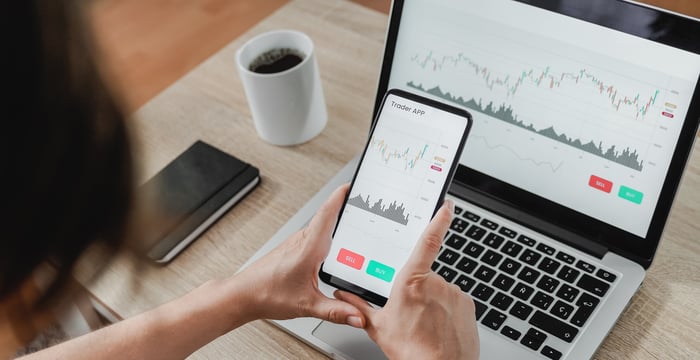 A person looks at a trading app on a phone and a laptop.