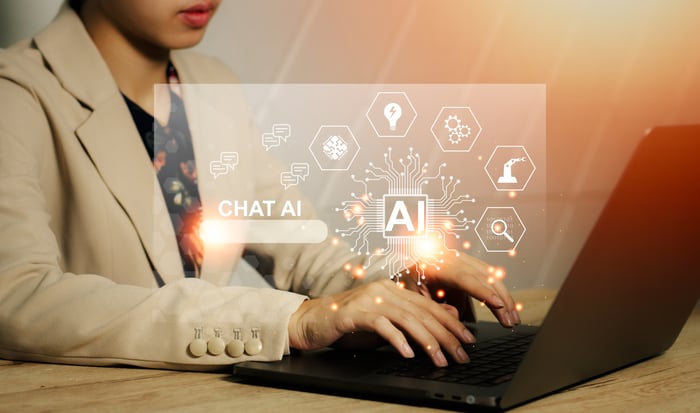 A person sits at a desk typing on the keyboard of a laptop computer as images of various icons, including AI, are overlayed on the photo