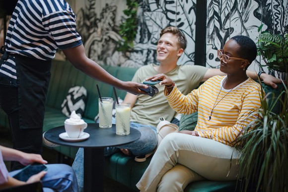 Three young friends sitting around a restaurant table while one is paying the tab using their phone.