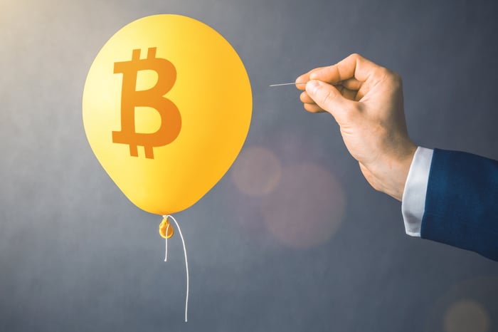 A balloon floating bears the Bitcoin logo as a hand holding a pin looms nearby, threatening to pop it.