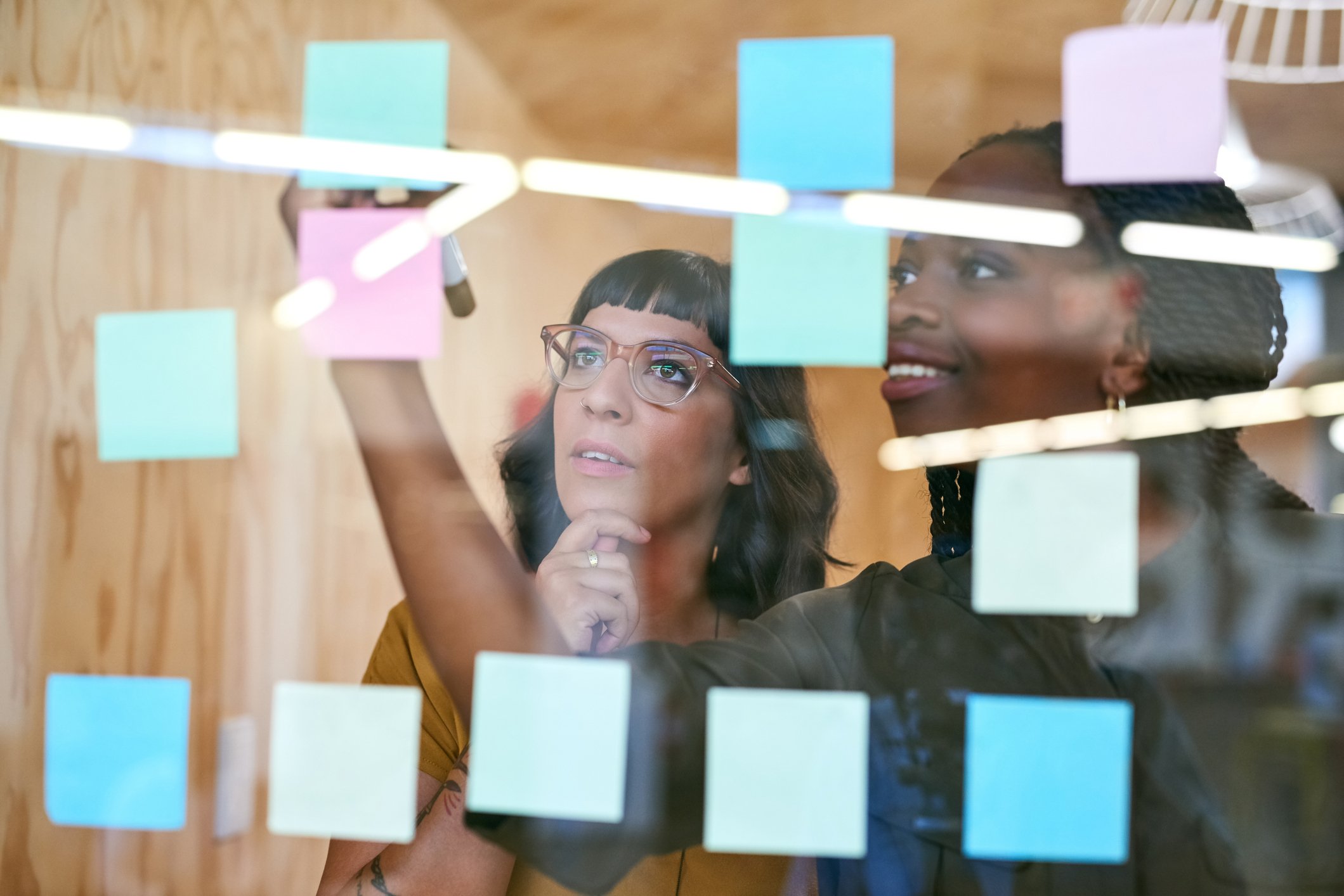 A pair of co-workers place sticky notes on a transparent office wall.