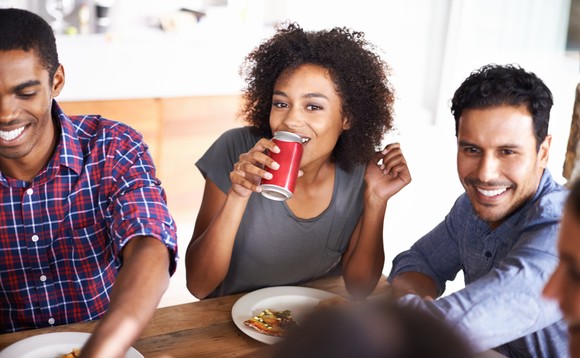 Five people sitting at a table while one drinks from a red can.