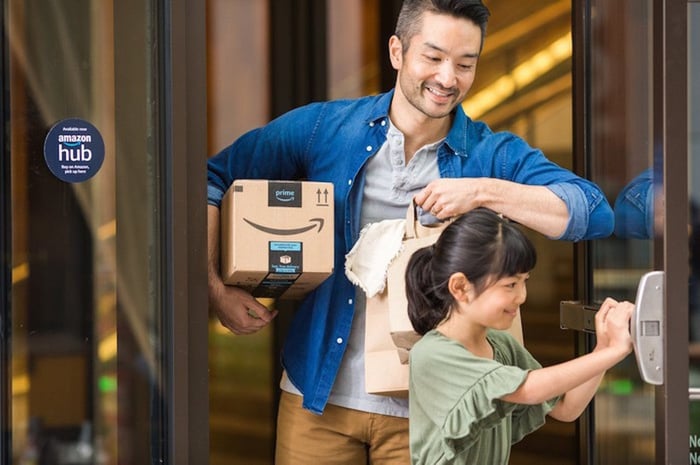 A parent carrying an Amazon package under their arm while their child holds a door open for them.