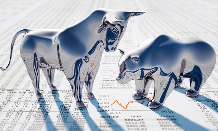 A silver bull figuring towers over a silver bear figurine, both standing on a newspaper showing stock prices.