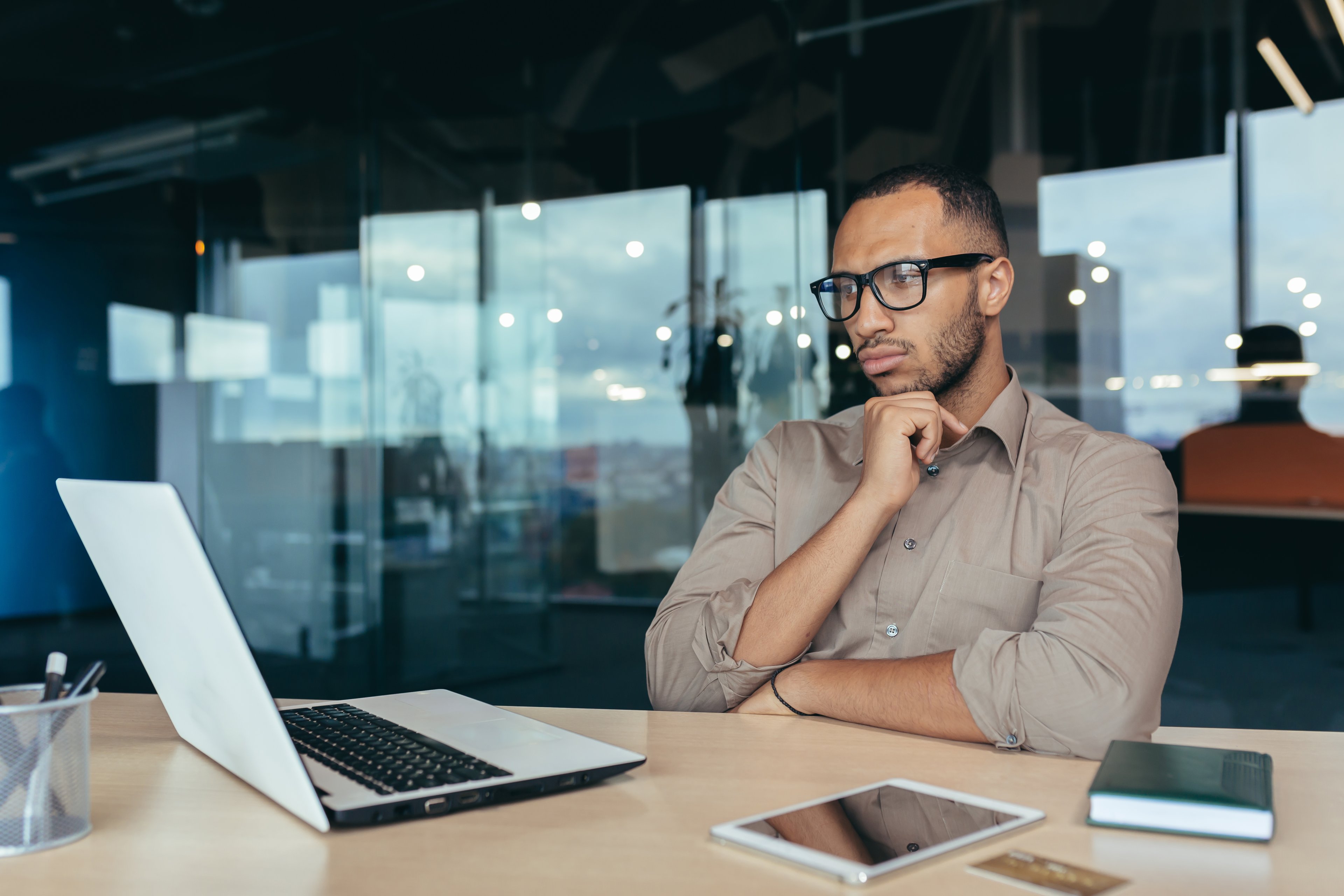 A person sitting at a desk, thoughtfully looking a at laptop in a modern office.