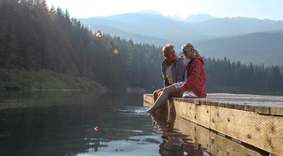 A couple sits on a dock in a mountain lake, dipping their feet in the water.
