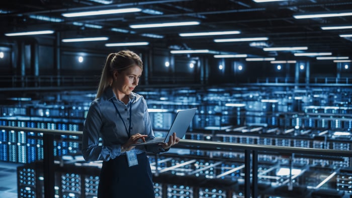 A person holding a laptop with servers in a data center in the background.