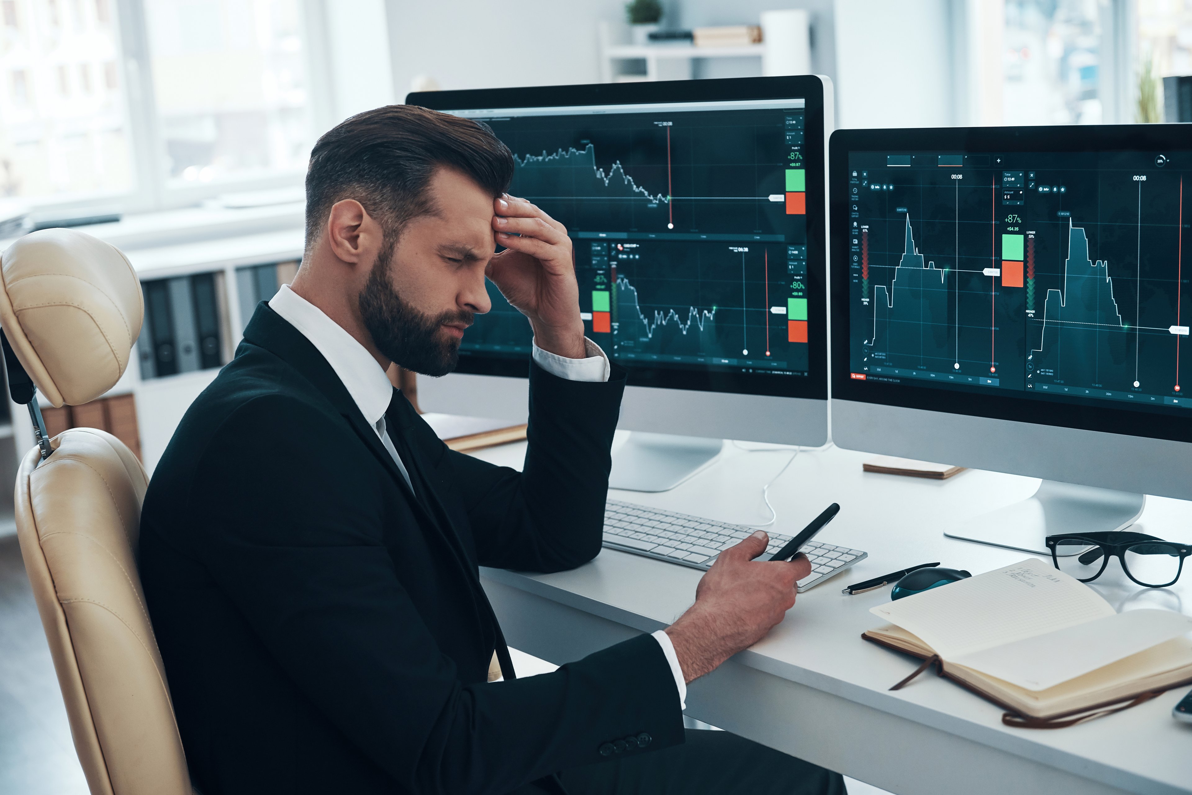 A person looking perplexed while seated at a desk in front of two computer screens displaying stock charts.