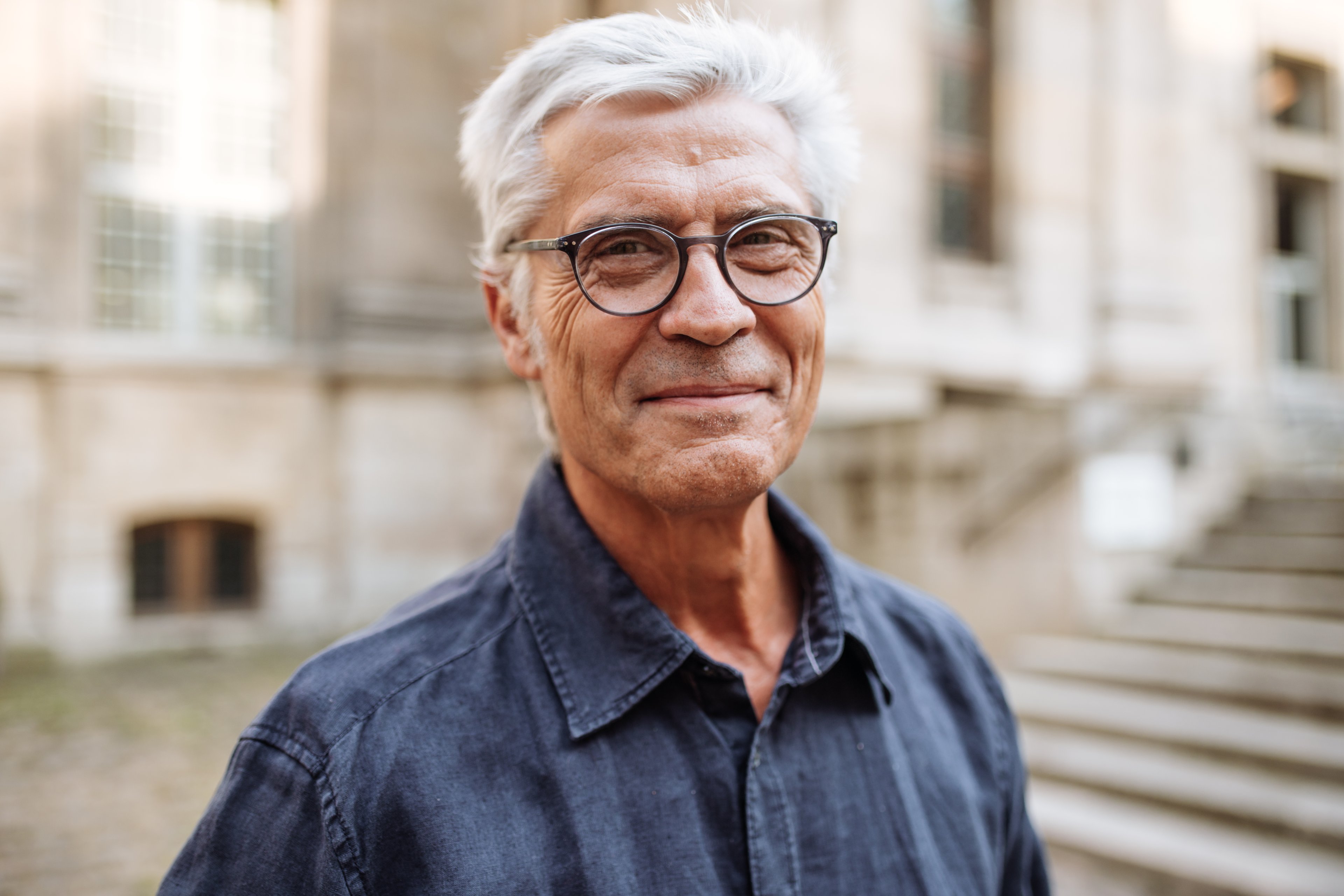 Someone with gray hair and glasses standing outdoors in front of a stone building.