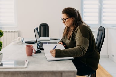 woman looks at laptop and writes in notebook.
