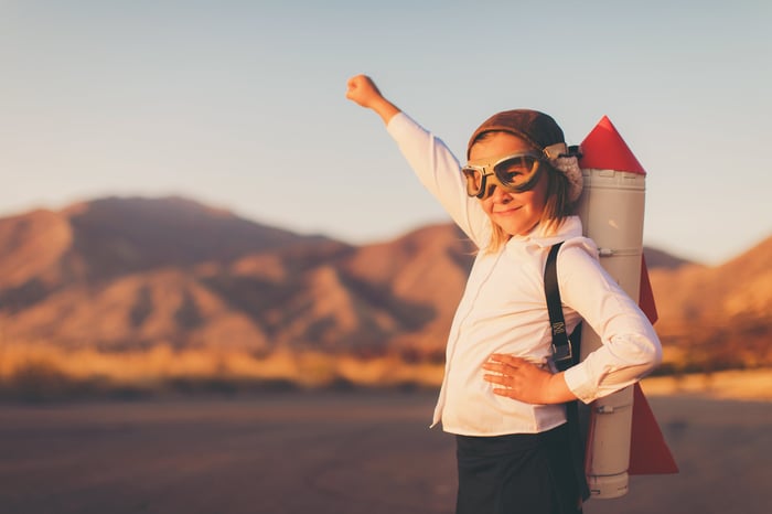 A kid with goggles and a toy rocket on the back, pointing toward the sky with fist. 