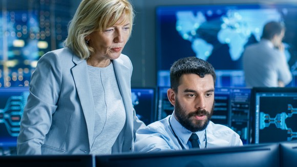Two people look at a computer screen in a high security command center. 
