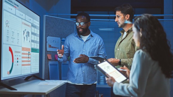 Three people standing around a computer screen and discussing a chart.