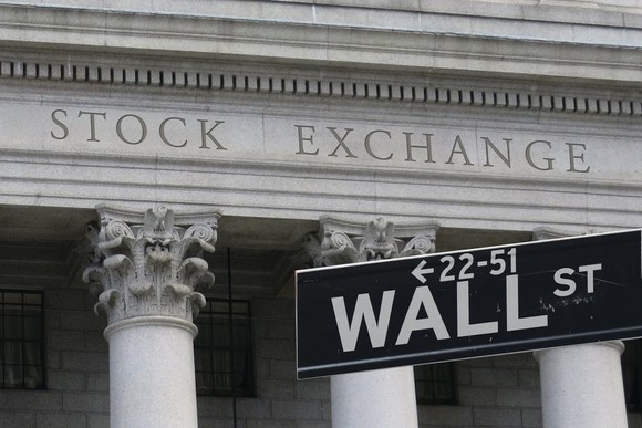 A stone building facade that says Stock Exchange behind a street sign for Wall St.
