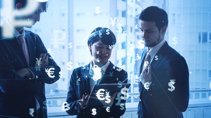 People looking at a tablet with digital currency symbols like dollar, euro, and yen floating around.
