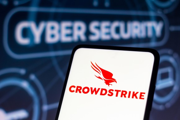 A red CrowdStrike logo sits against white background on a phone, which is being held up to a blue and electronic-looking backdrop that says Cyber Security.