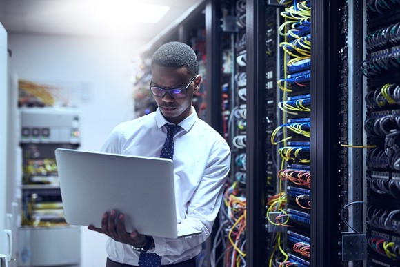 An engineer using a computer while standing next to a row of servers.