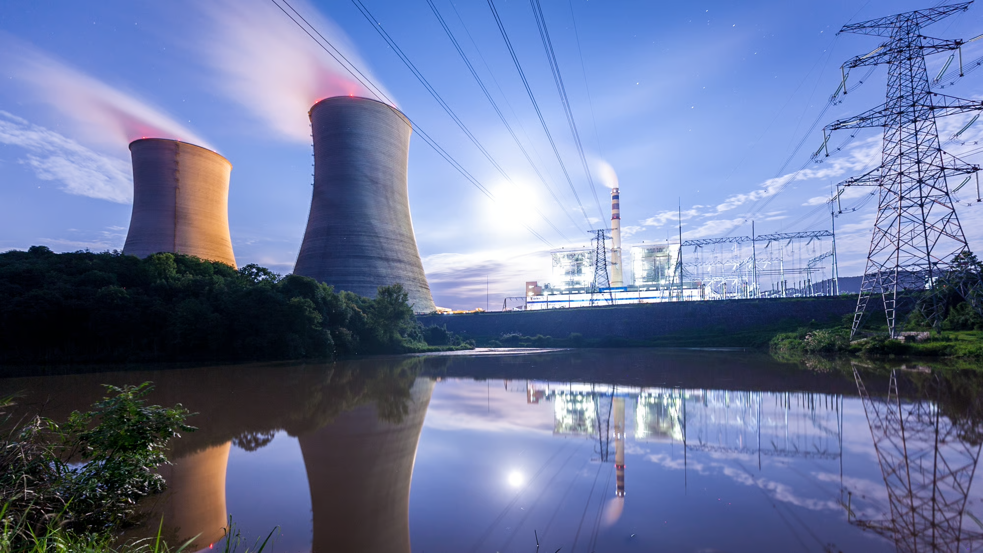 Two nuclear reactors at twilight. In the foreground, a small pond reflects the reactors and some powerlines. 