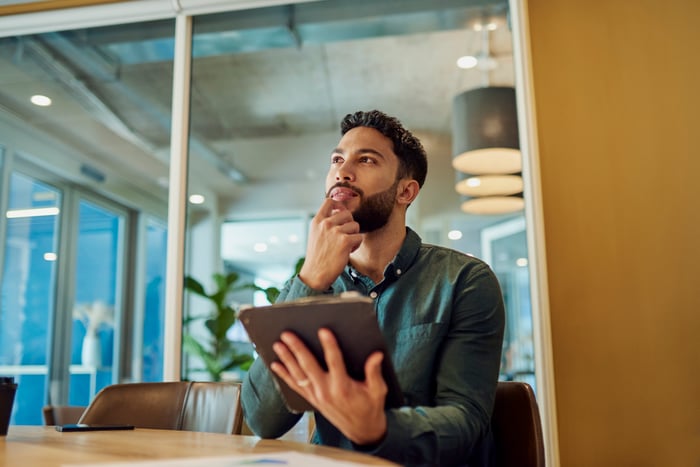 A person sitting at a table holding a tablet, appearing deep in thought while looking upward.