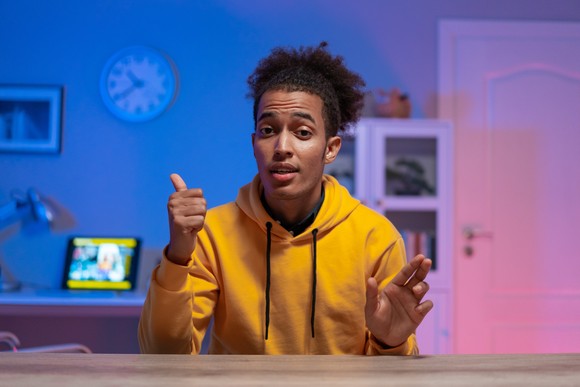 Person in yellow hoodie use thumb to point at clock in the background.