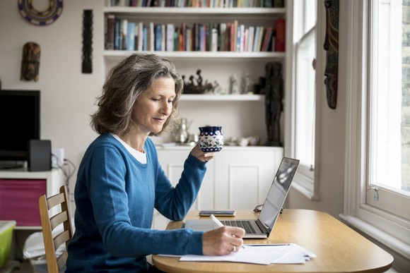 A person sits at a desk holding a mug in one hand and making a note on paper with the other.