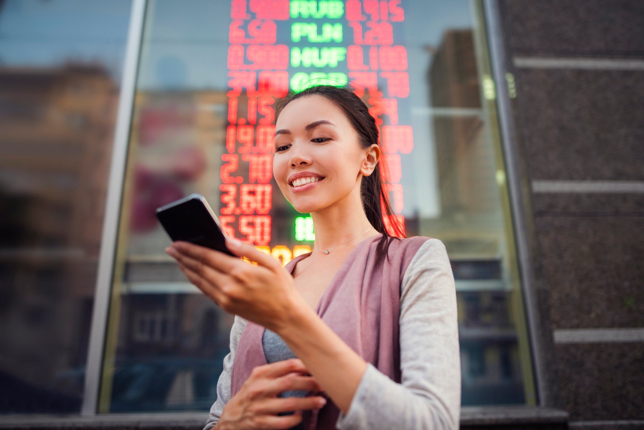 An investor stands in front of an electronic panel in a city and looks at something on a smartphone.