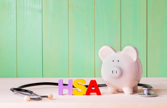 On a table in front of a pale green wall are a stethoscope, block letters spelling out HSA, and a pink piggy bank.
