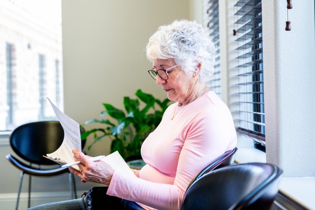 An older woman sits in an office chair, looking through paperwork. 